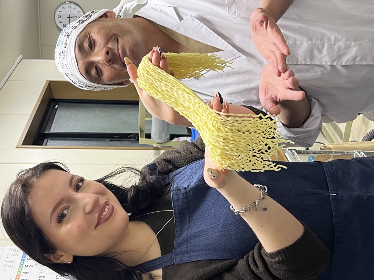 A guest proudly holding up her freshly cut wavy noodles next to a smiling Taka