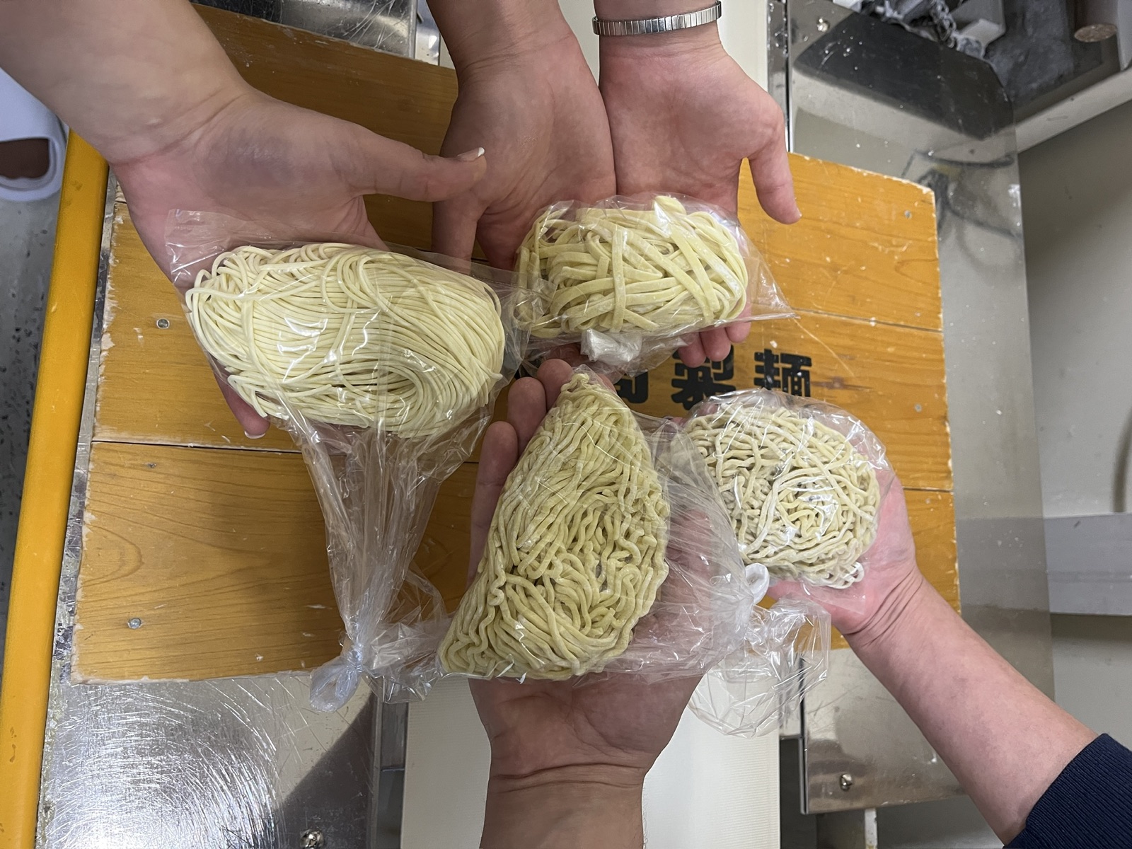 Four hands holding four bags of fresh noodles in different thicknesses and shapes